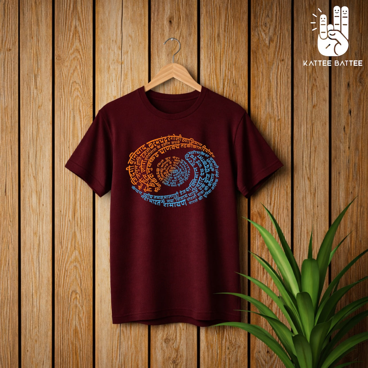 A maroon short-sleeve t-shirt hanging on a wooden hanger against a vertical wood plank wall, featuring a colorful spiral graphic made of script that depicts Doordarshan Nostalgia. A green plant is visible in the lower right corner, and the KATTEE BATTEE logo is in the top right.
