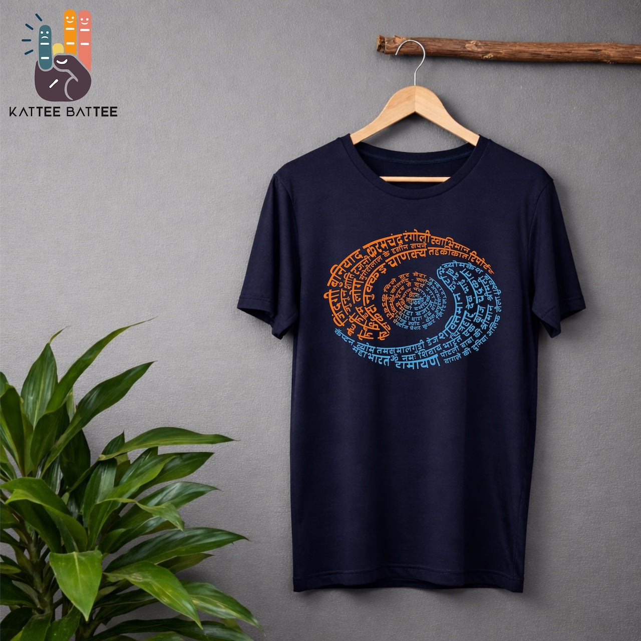 A navy blue short-sleeve t-shirt hanging on a wooden hanger against a gray wall, featuring a circular mandala-style yin yang like graphic made of orange and blue Devanagari script that depicts Doordarshan Nostalgia. A green plant is visible on the left, and a wooden branch hangs above the shirt.